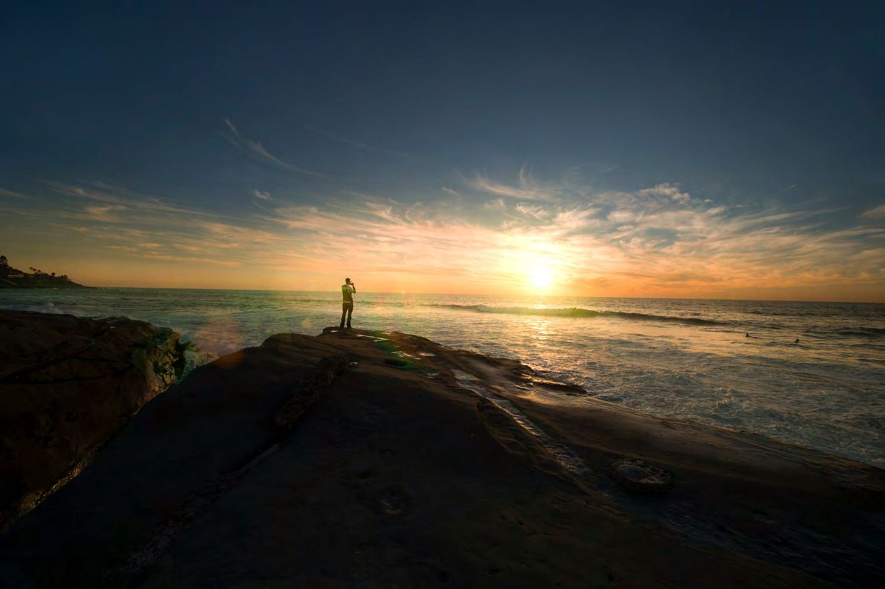 Person stands on rocky shore at sunset.